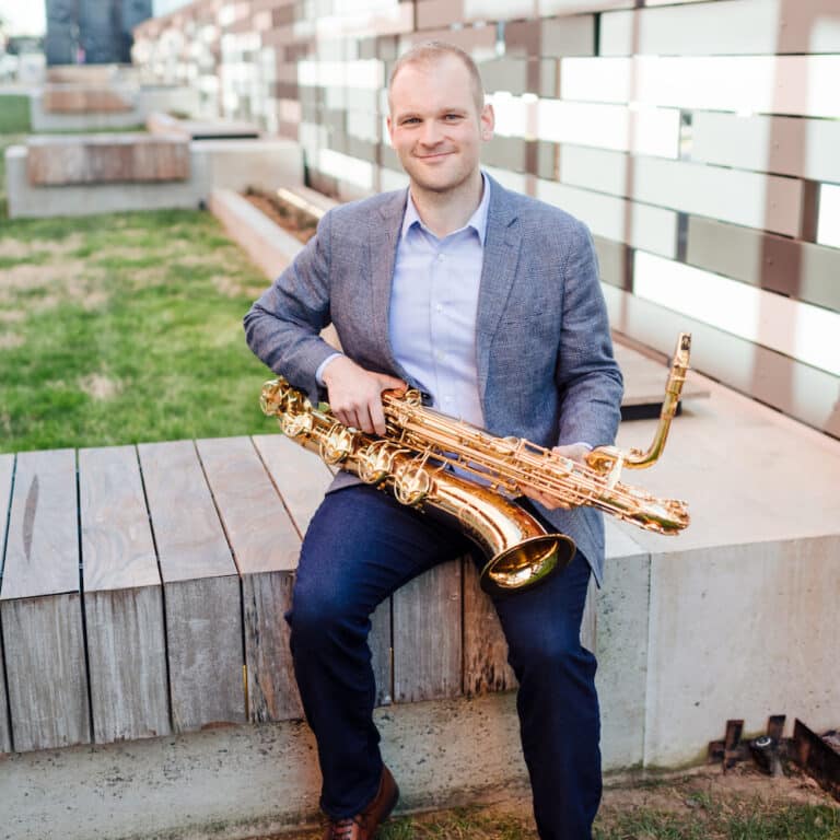 Ryan Middagh sits on a concrete bench outside, smiling into the camera and holding his saxophone.