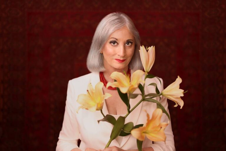 Renee Rosnes looks directly into the camera against a dark red background, foregrounded by some large yellow flowers.
