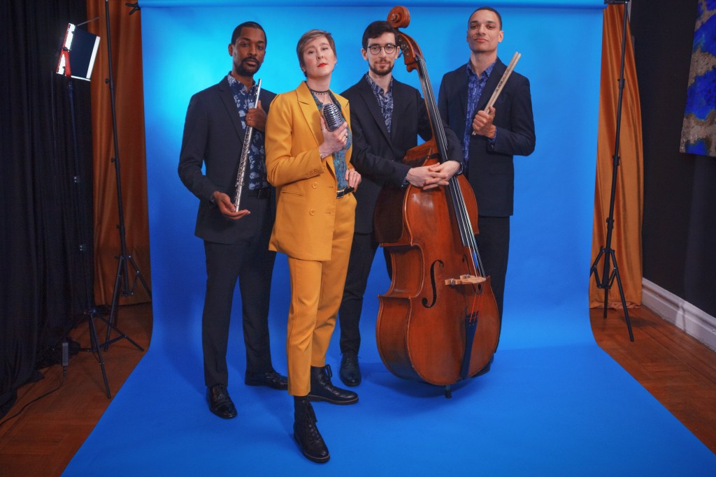 The quartet stand in a row, in a studio against a blue background. They each carry their instrument or something to do with it; Thomas with a flute, Green with a microphone, Rosenfeld with his double bass and Hyde with his drumsticks. They look directly into the camera.