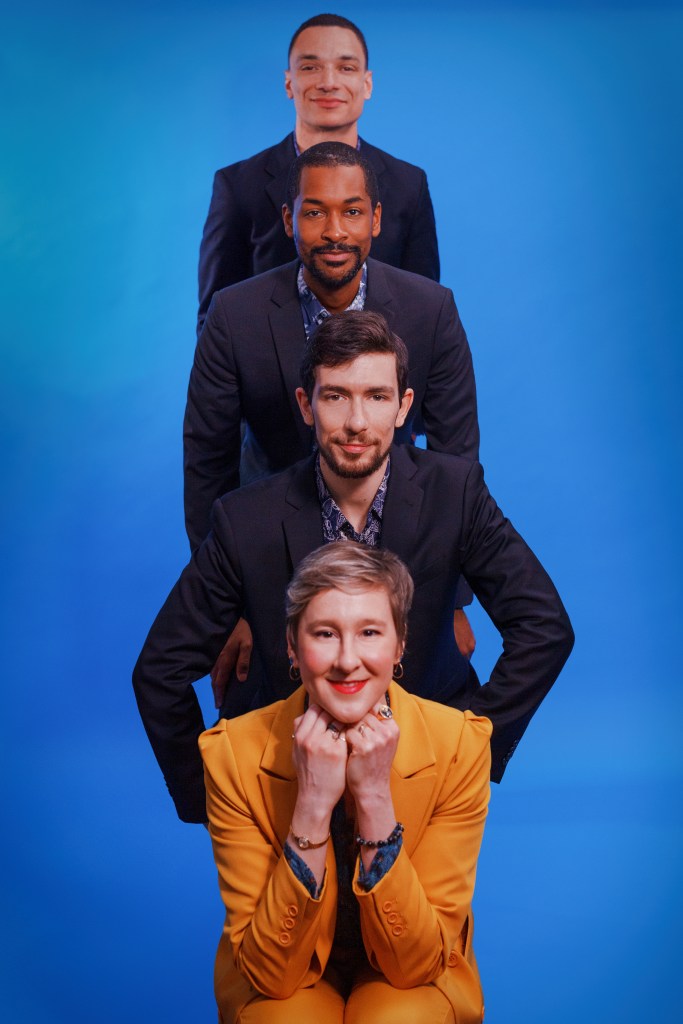 The quartet are positioned so their heads are on top of one another in a vertical row, against a blue background, with Green in the front. They smile into the camera.