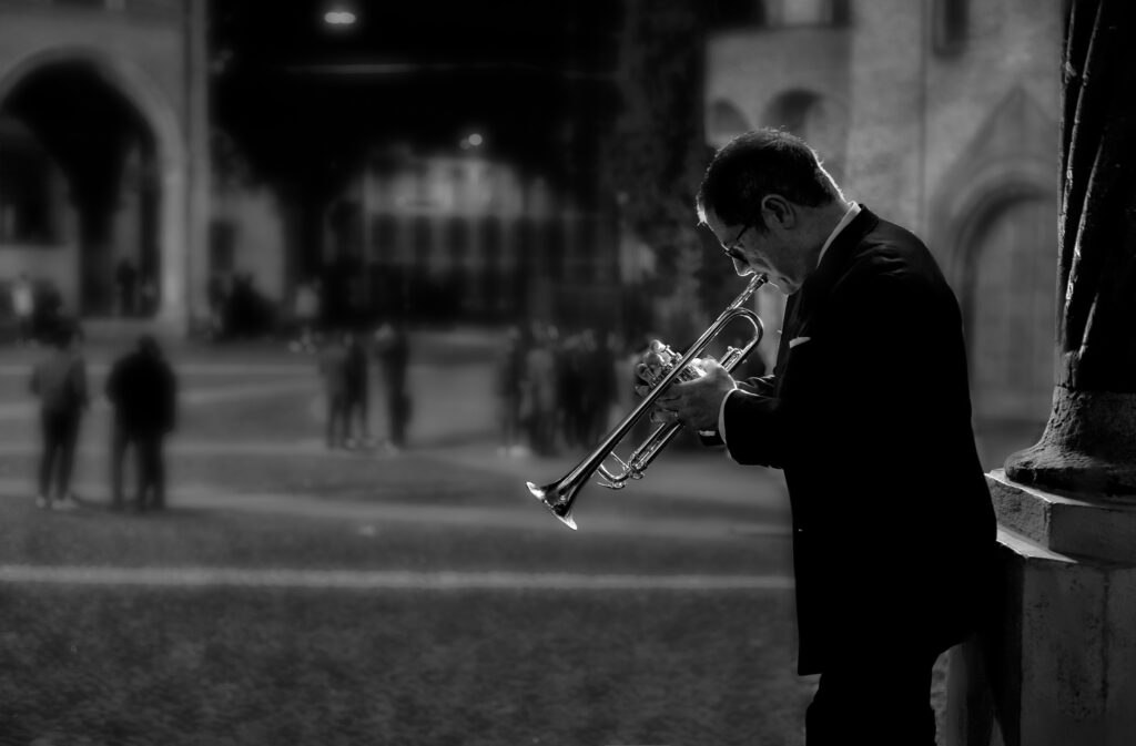 Black and white picture. Jim Rotundi plays the trumpet in side profile, against a blurred backdrop of a pretty square filled with people and old buildings. He is wearing a suit and leans against a stone pillar.