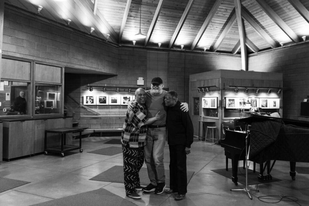 The three musicians stand together in a recording studio with their arms around each other, smiling.