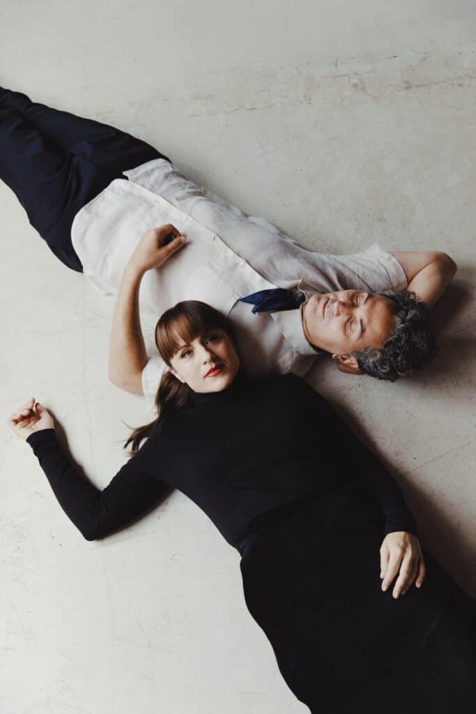 Rachel Eckroth and John Hadfield lie on the white concrete ground. Rachel is looking up at the camera and John has his eyes closed.