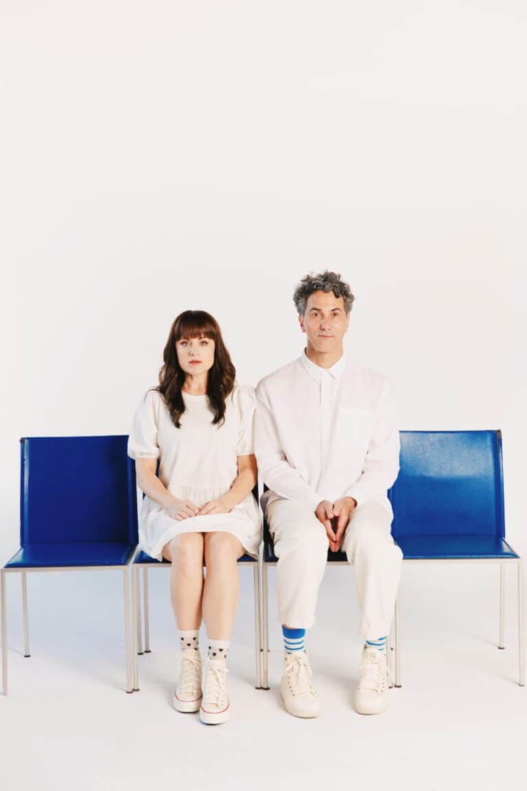 The two musicians sit on a blue bench against a plain white background. They are both wearing all white and looking straight into the camera.