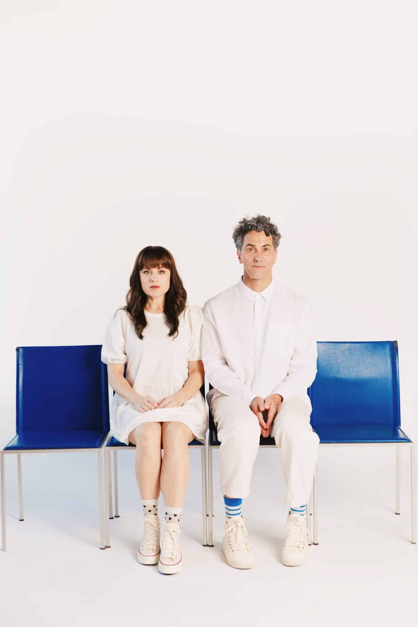 The two musicians sit on a blue bench against a plain white background. They are both wearing all white and looking straight into the camera.