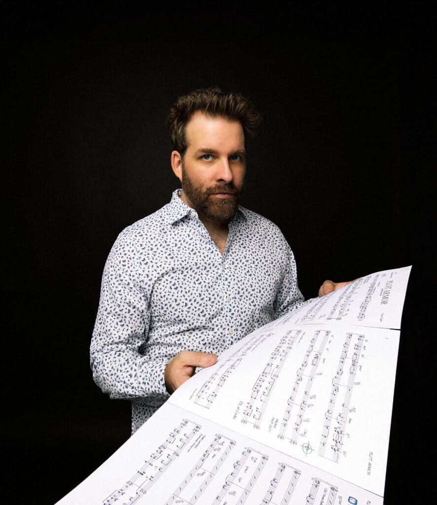 Taylor Eigsti stands against a black background, looking straight into the camera. He holds sheets of music.