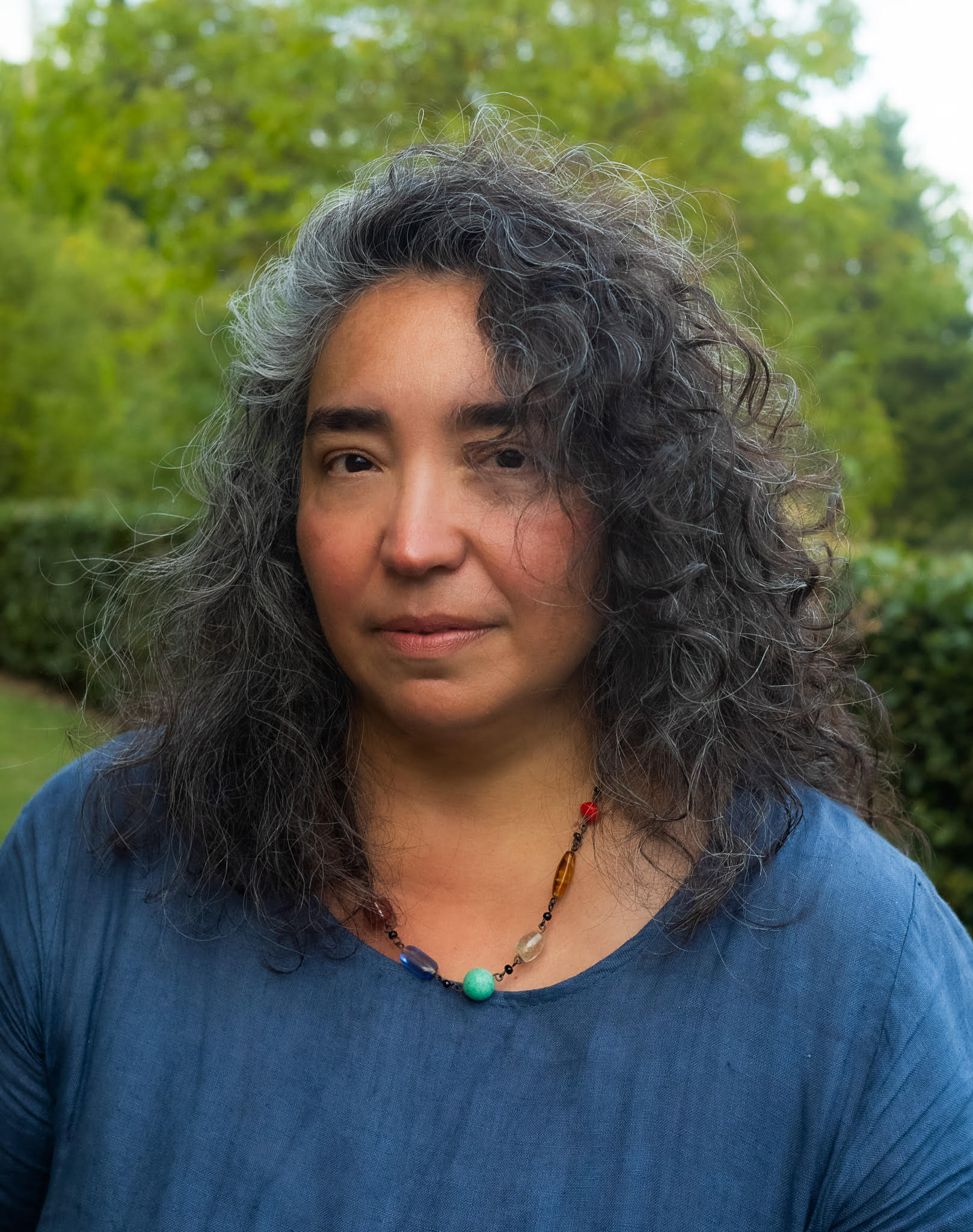 Angelica Sanchez headshot outside with greenery in background. Wearing blue top and colourful necklace