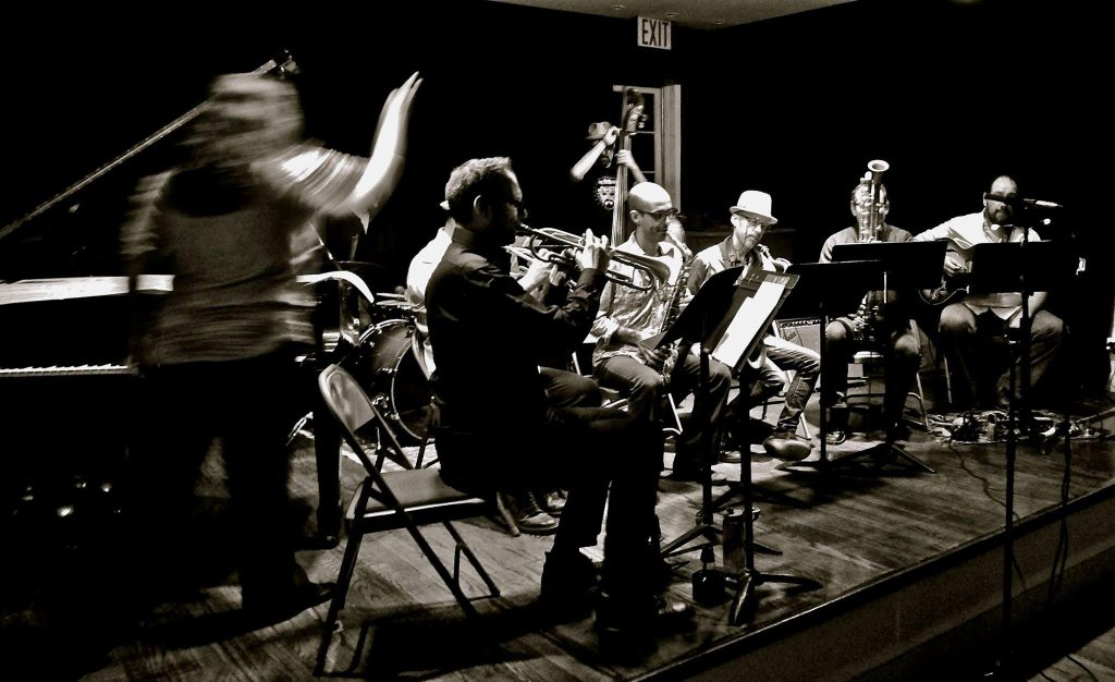 Angelica Sanchez conducting her nonet on stage from the piano. Image in black and white with blurry movement