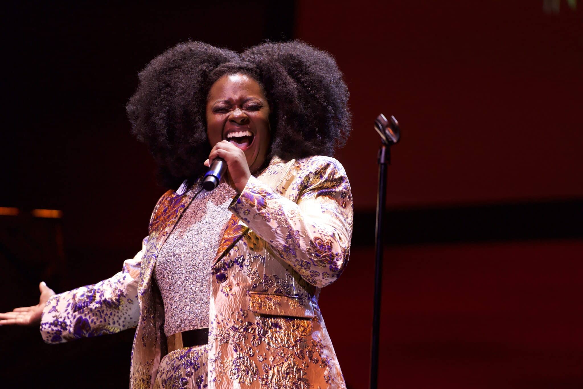 April May Webb stands on stage in a shiny patterned suit, singing passionately into the microphone.