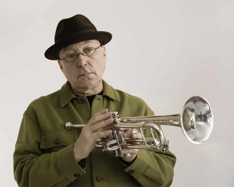 Brian Lynch stands against a white background, looking off to the side and holding his trumpet.