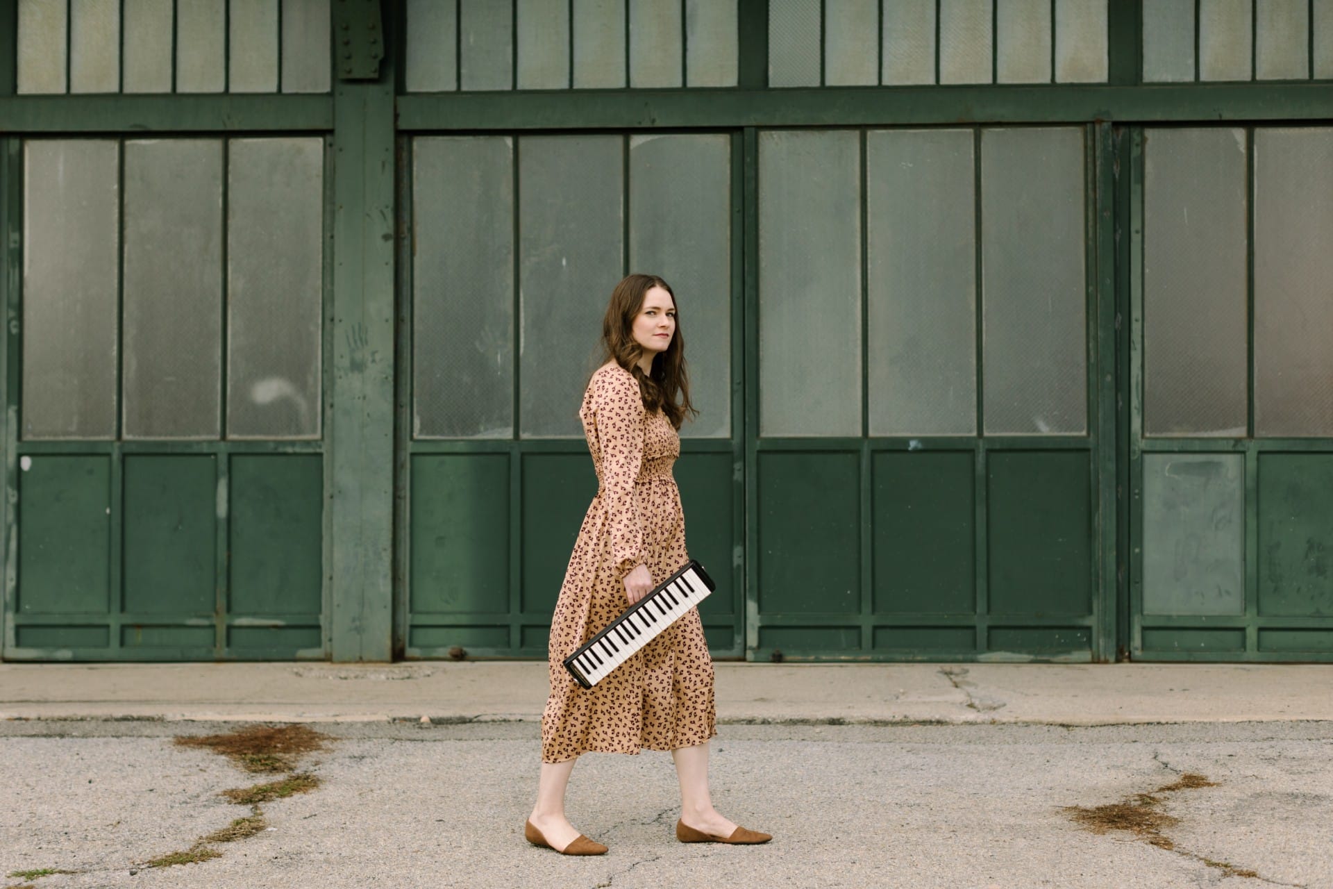 Caili O'Doherty walks along a concrete street, carrying a mini keyboard and looking sideways to camera.