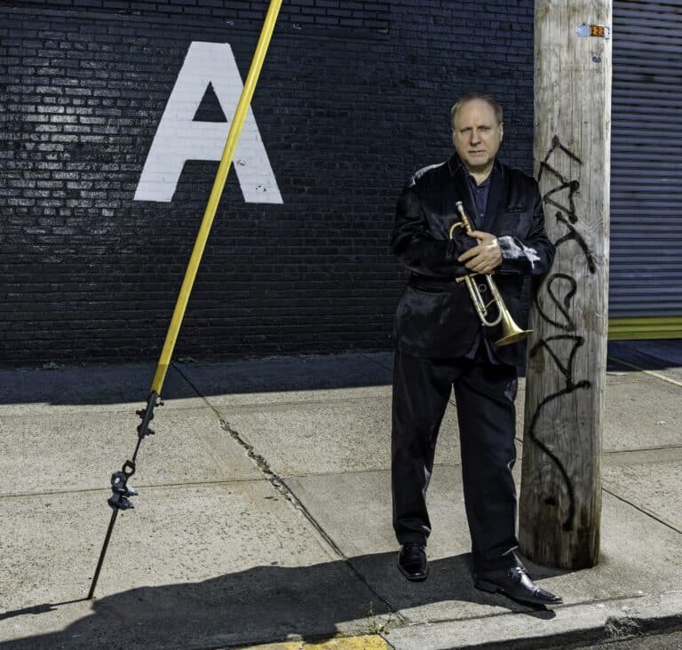 David Weiss stands on a sunny sidewalk, holding his trumpet and looking directly into the camera. A yellow cable cuts across the picture to the left, and a large letter 'A' is painted on the wall behind him.