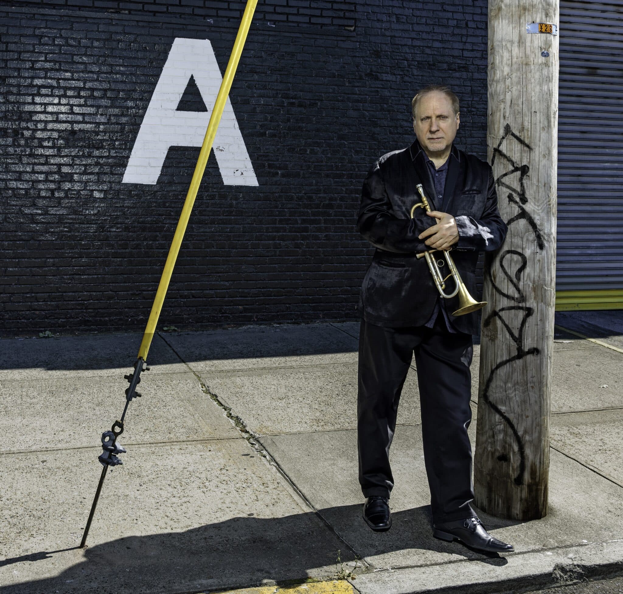 David Weiss stands on a sunny sidewalk, holding his trumpet and looking directly into the camera. A yellow cable cuts across the picture to the left, and a large letter 'A' is painted on the wall behind him.