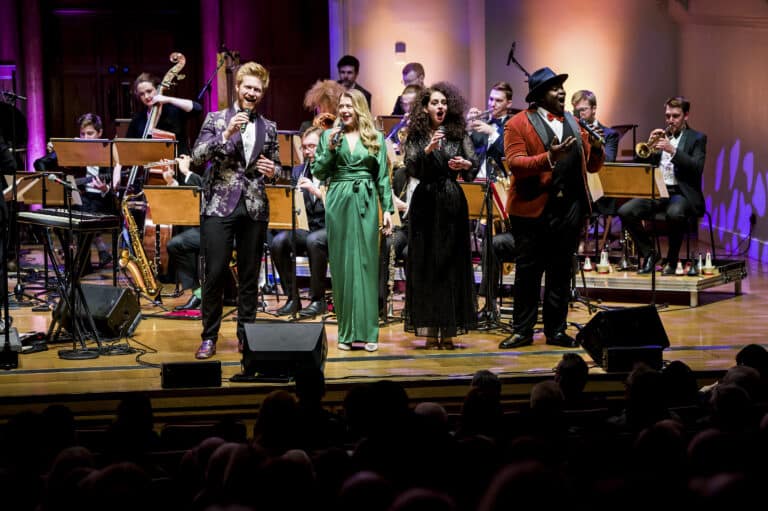 Four singers stand at the front of the stage with mics, looking jubilant mid-song. The rest of the orchestra is behind them.