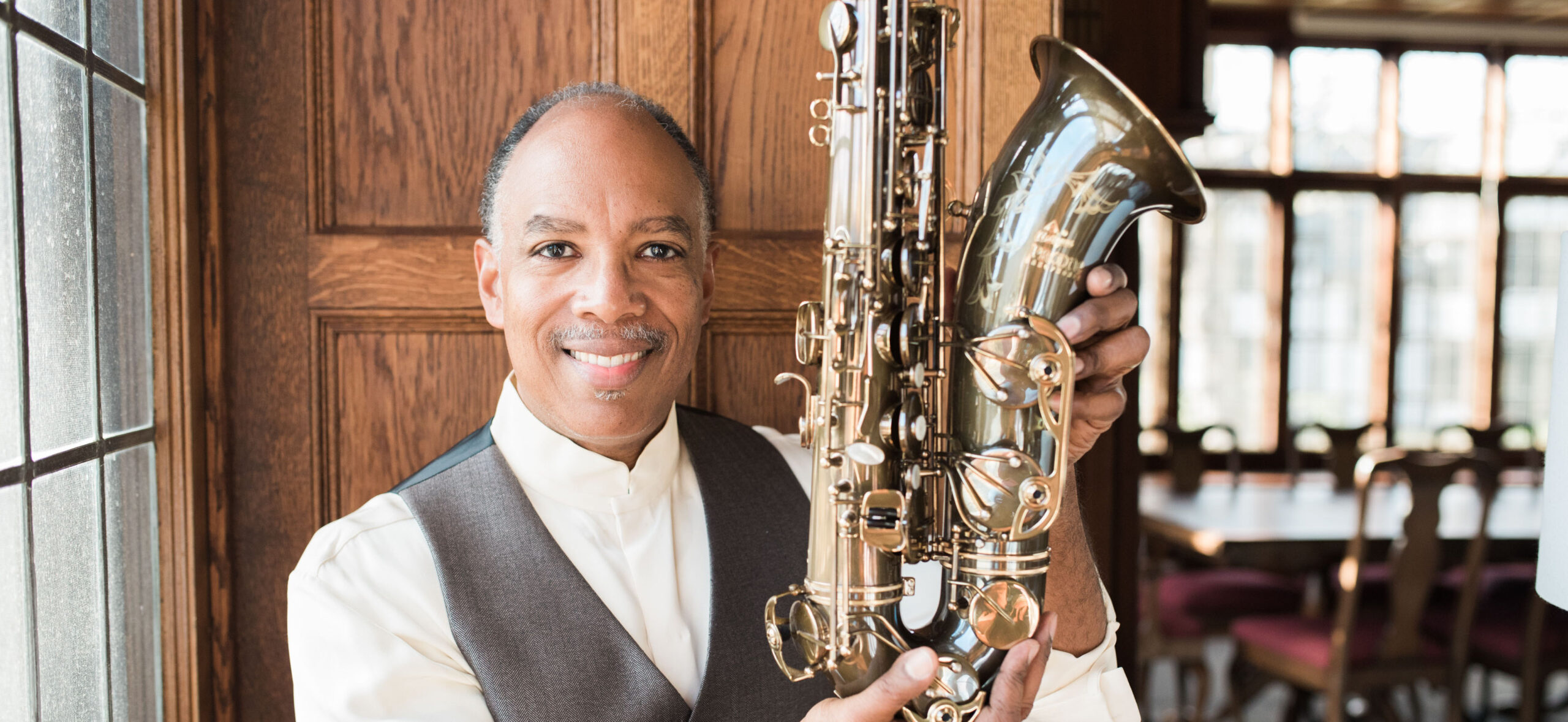 Don Braden sitting and smiling holding his saxophone up to he camera, wearing white shirt and grey waistcoat. Dark wood panelling and windows in the background