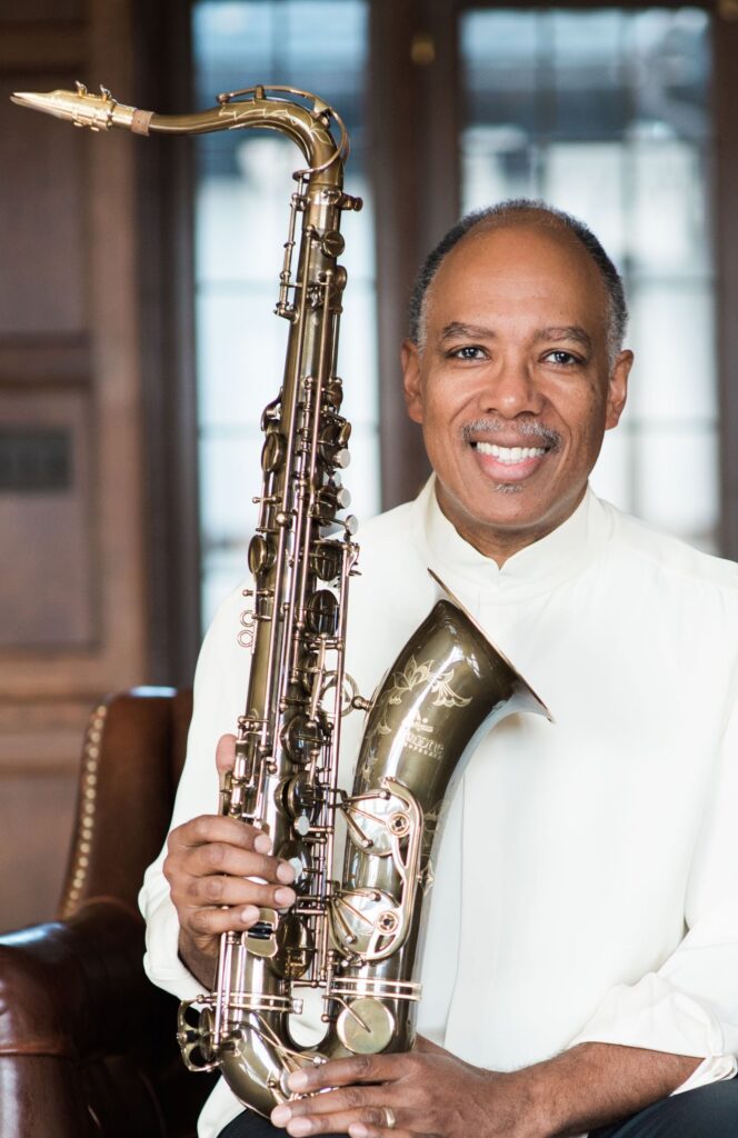 Don Braden sitting in a chair wearing a white shirt, smiling and holding his saxophone