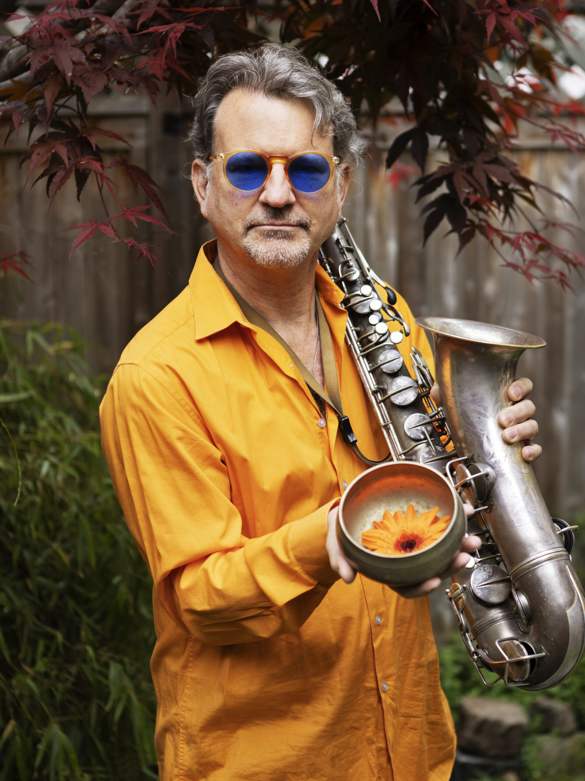 Jeff Lederer outside holding tenor sax and an orange flower, wearing sunglasses and an orange shirt