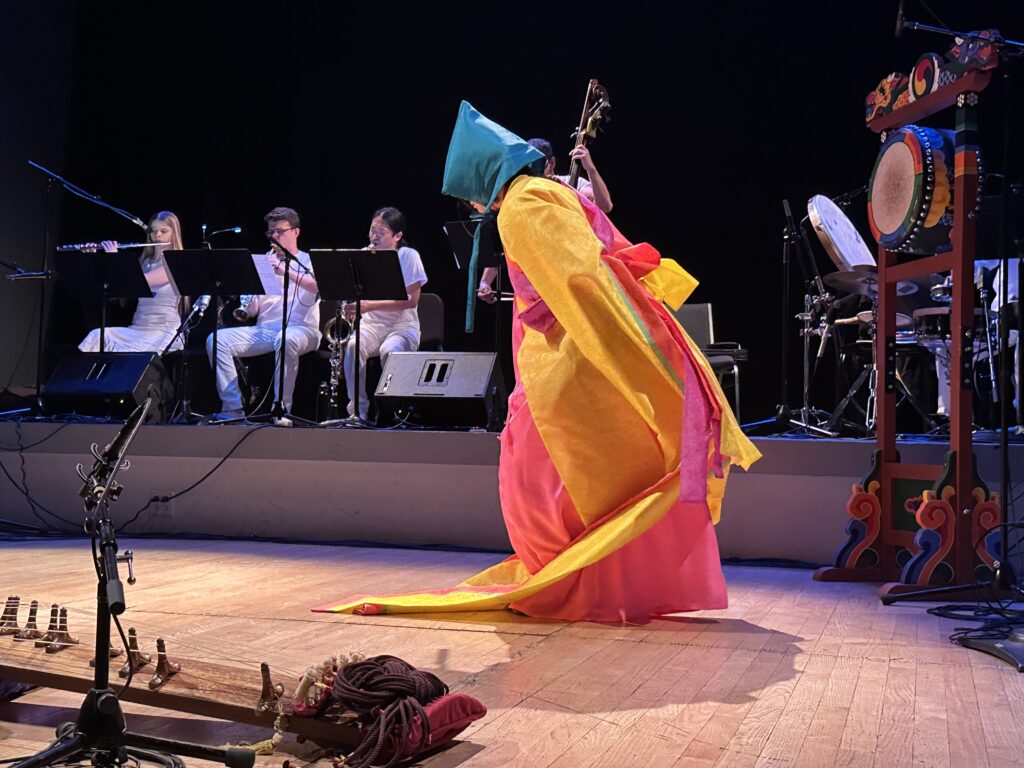 DoYeon Kim dancing on stage wearing bright yellow and pink robes with a turquoise coloured hood covering her head and face. Musicians in the background