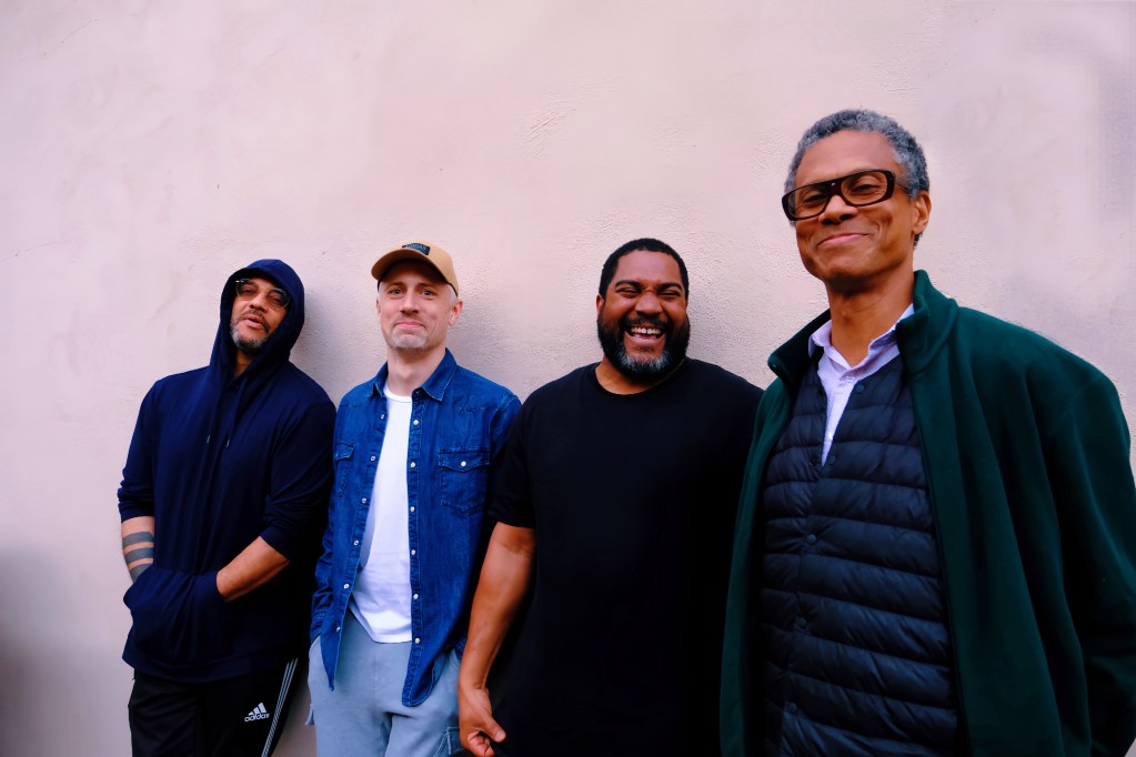 Four musicians stand in a row against a white wall, looking directly into the camera and smiling.