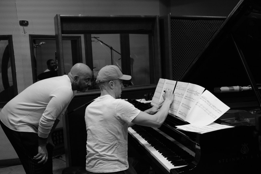Black and white photo. Eric Revis looks over John Escreet's shoulder, who is sitting at the piano making notes on sheet music.