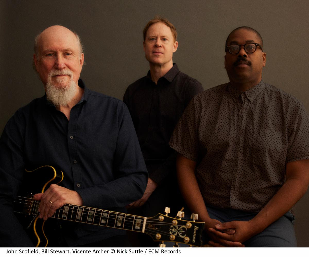 John Scofield, Bill Stewart and Vicente Archer all wearing black and facing the camera with a lighter black background