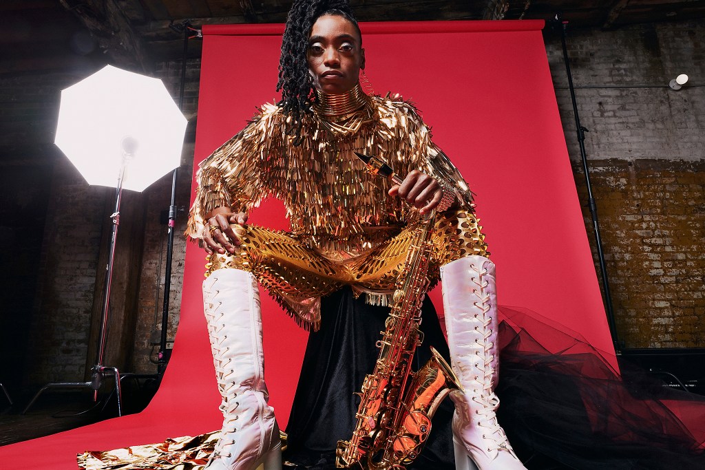 Lakecia Benjamin wears a gold sparkly top and trousers, and knee high white boots. She looks down at the camera holding her saxophone.