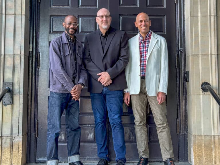 The three musicians stand side by side in front of a large wooden door, smiling into the camera.