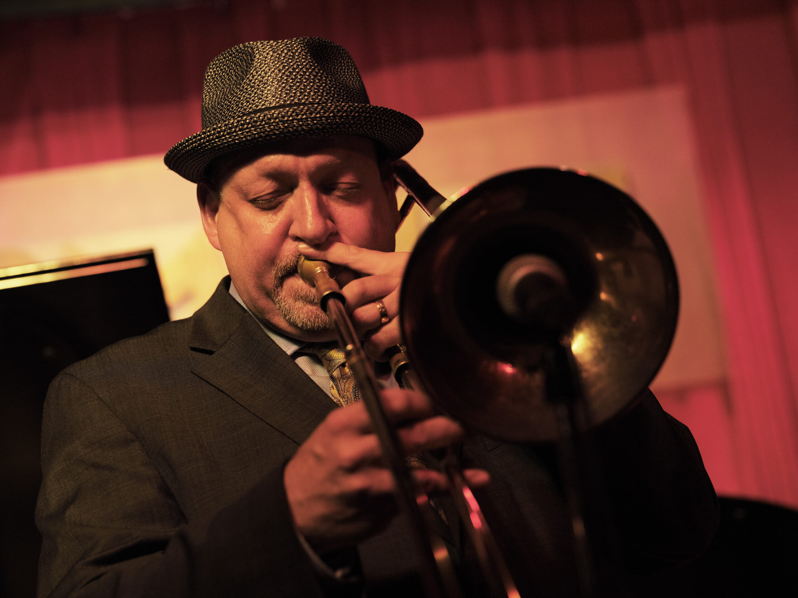 Steve Davis playing trombone. He wears a grey fedora and has his eyes closed as he blows the instrument, against a warmly lit background.