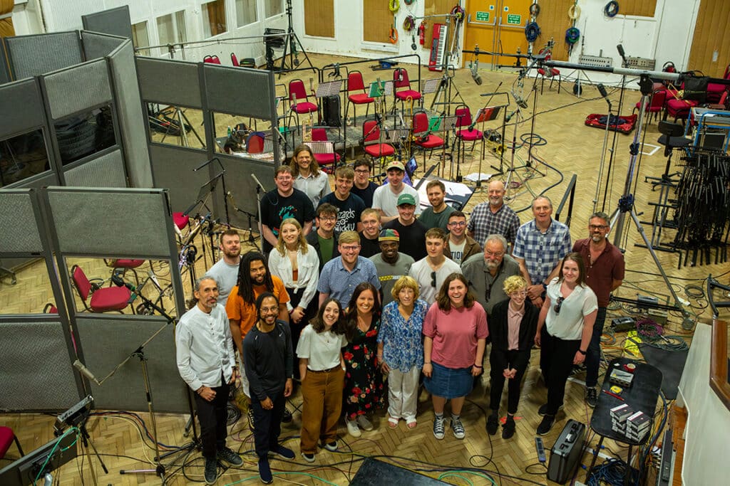 Aerial shot of a large group of musicians, standing in a group in a recording studio, looking up at the camera and smiling.