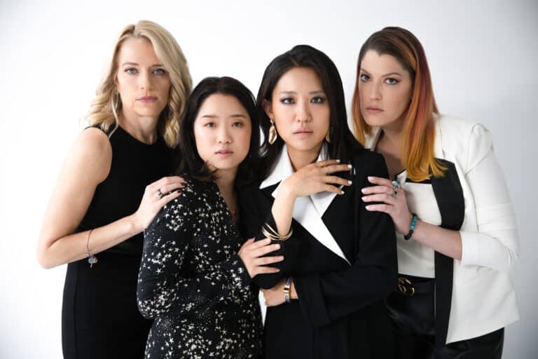 The four women, dressed elegantly in black and white, stand looking straight into camera with their hands on each other's shoulders.