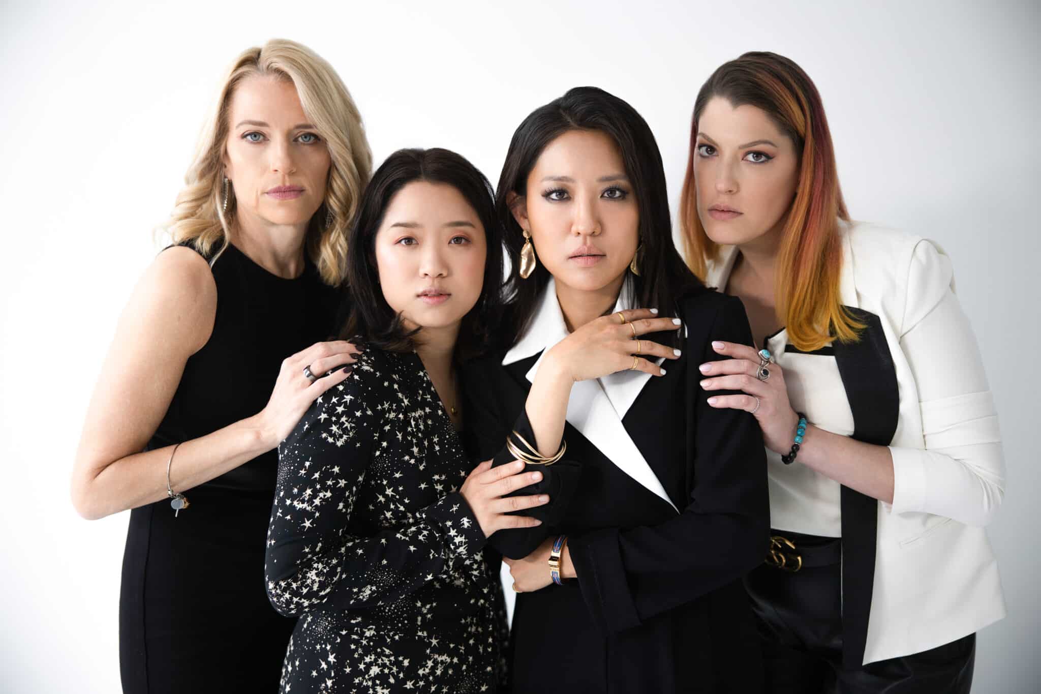 The four women, dressed elegantly in black and white, stand looking straight into camera with their hands on each other's shoulders.