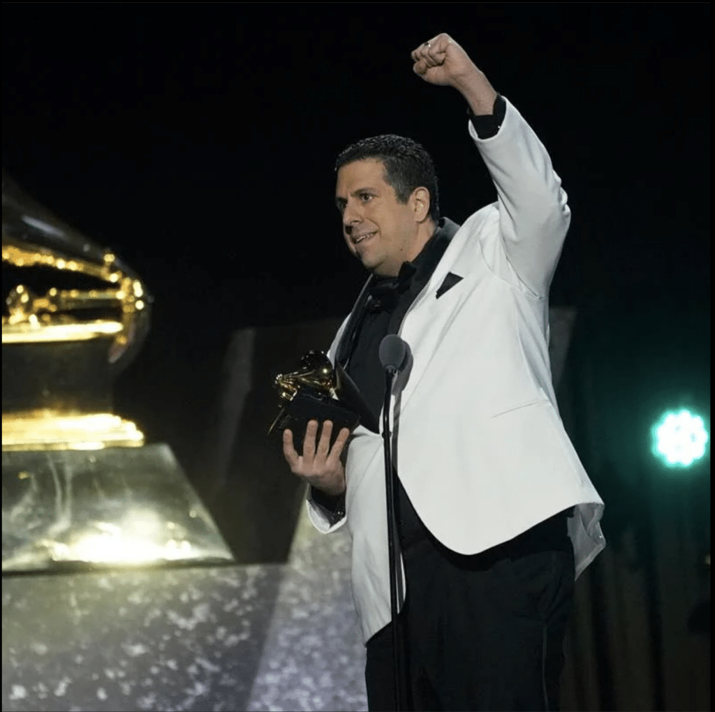 Ricky Riccardi stands on stage at the Grammys holding his award, and pumping his fist in the air.
