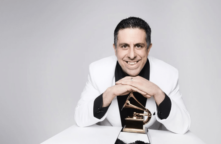 Ricky Riccardi sits in a white studio in a white suit, with his hands folded over his Grammy award, smiling into camera.