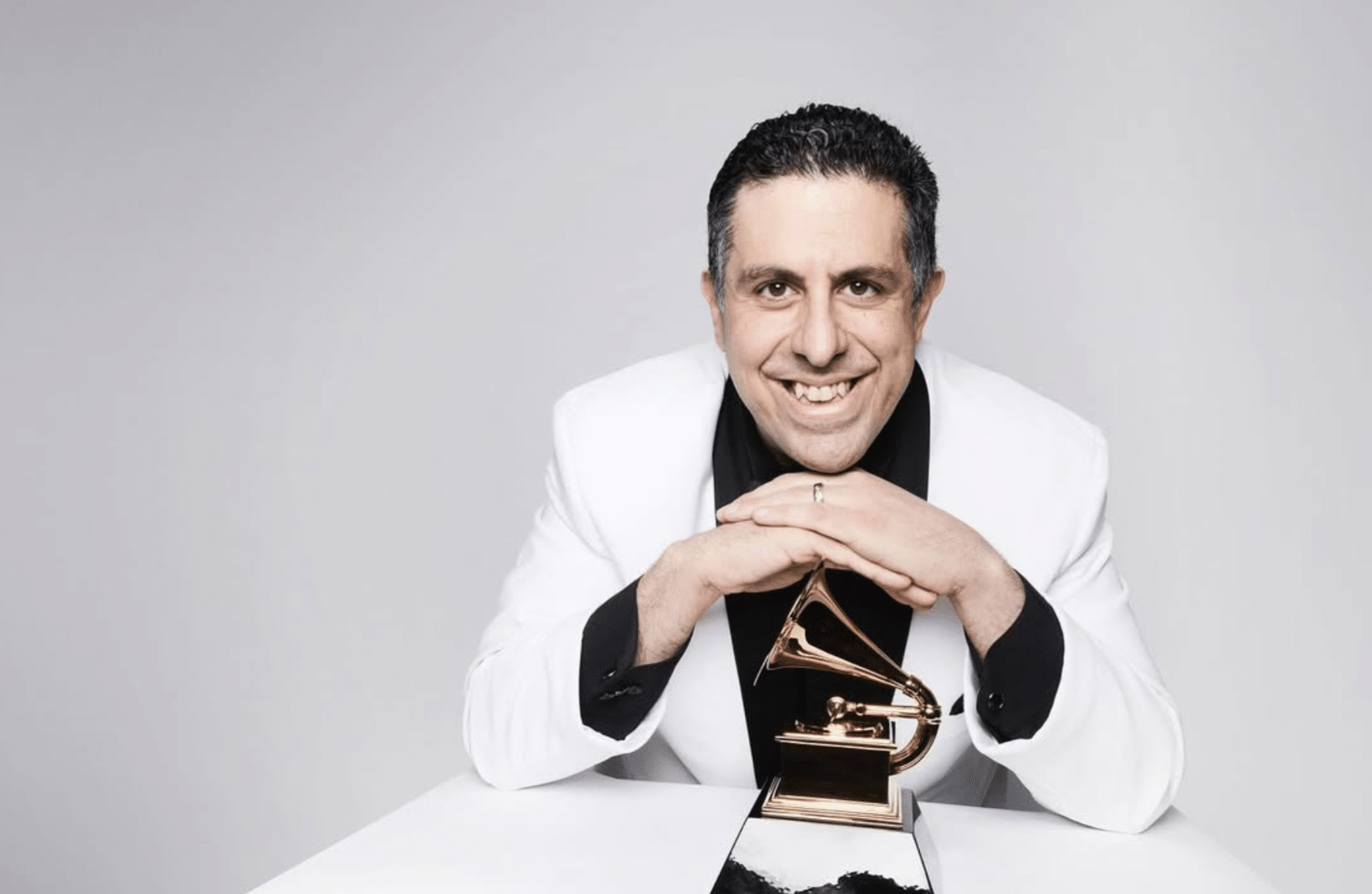 Ricky Riccardi sits in a white studio in a white suit, with his hands folded over his Grammy award, smiling into camera.