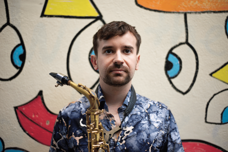 Head and shoulders shot of Tom Smith, holding his saxophone and looking slightly off camera. The background is a white wall with colourful geometric art on it.